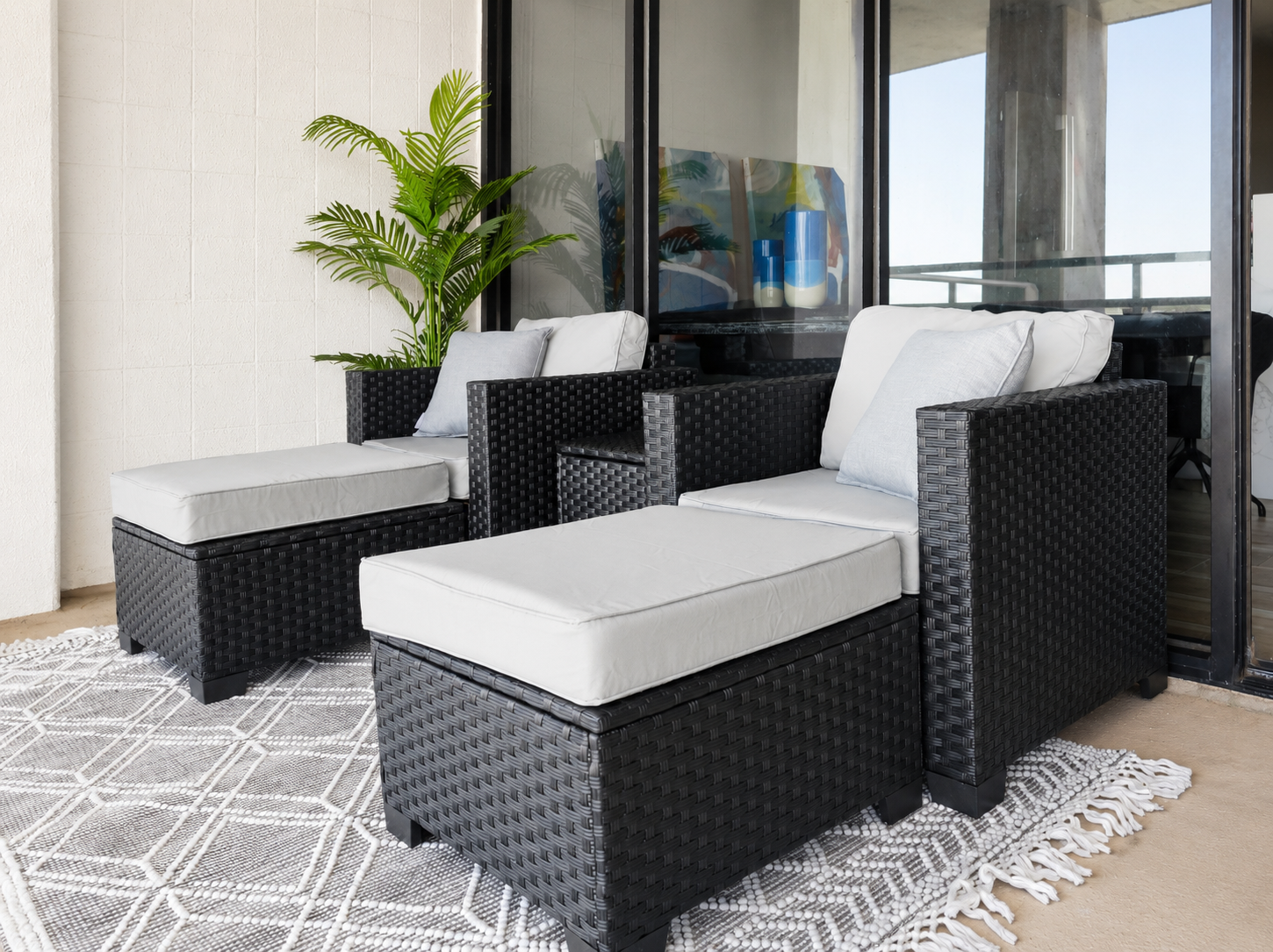 Modern coastal balcony with black wicker lounge chairs, light gray cushions, matching ottomans, geometric outdoor rug, and contemporary condo patio overlooking the ocean