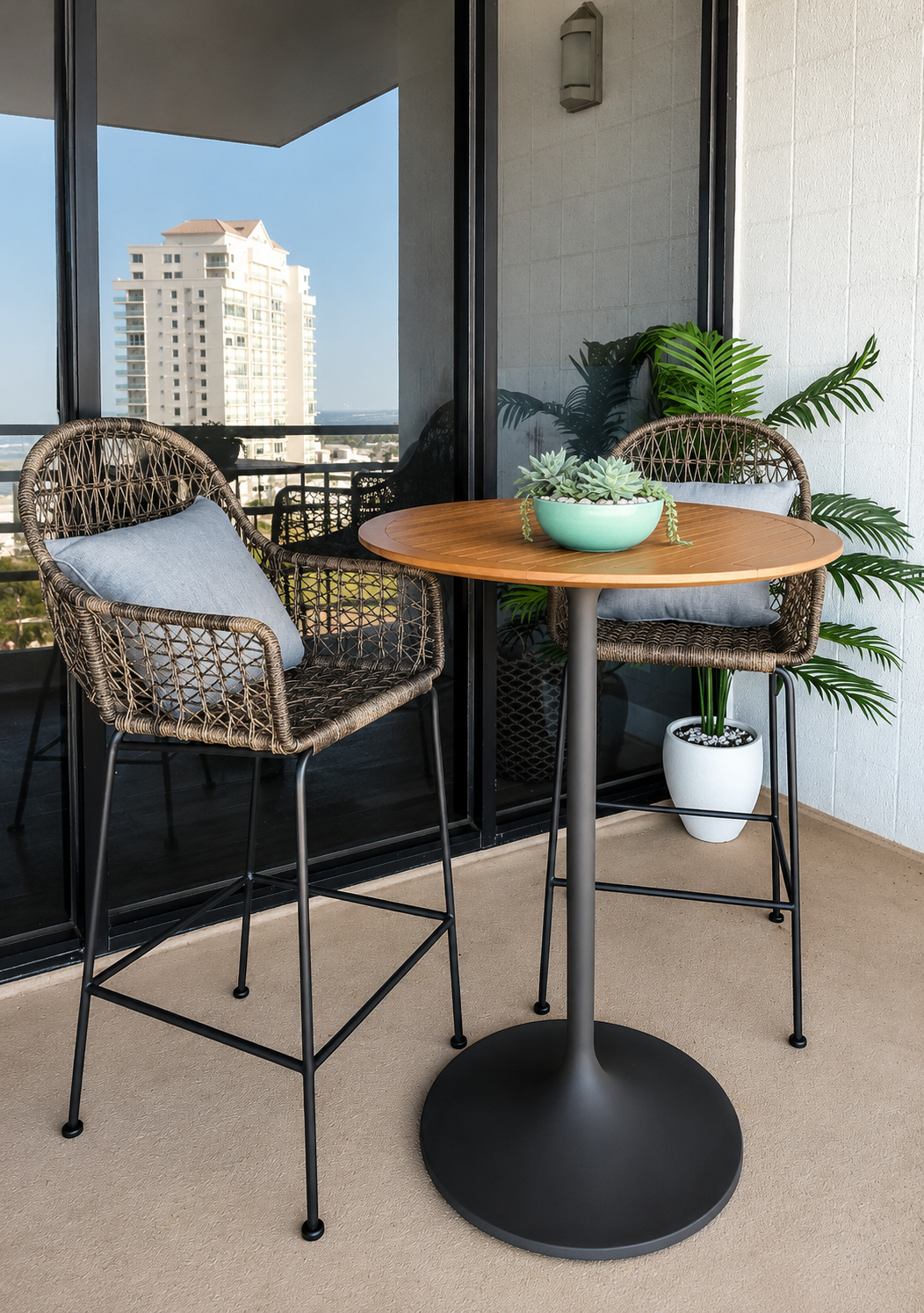 Modern coastal condo balcony with woven bar-height chairs, round wood-top café table, lush potted greenery, and contemporary outdoor seating overlooking the ocean