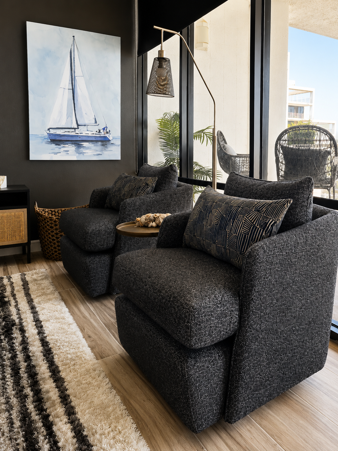 Modern coastal living room with textured charcoal lounge chairs, black accent wall, sailboat artwork, floor-to-ceiling windows, and balcony views in a luxury waterfront condominium