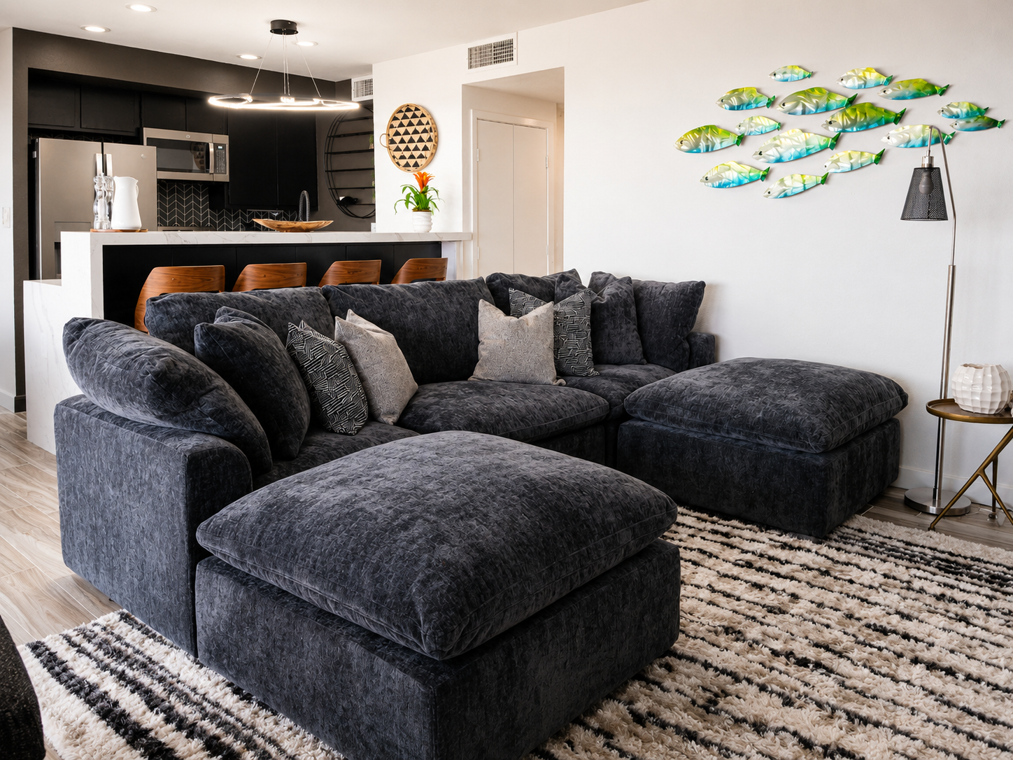Modern coastal living room with a charcoal sectional sofa, black kitchen with waterfall island and wood bar stools, contemporary fish wall art, and luxury open-concept condominium interior