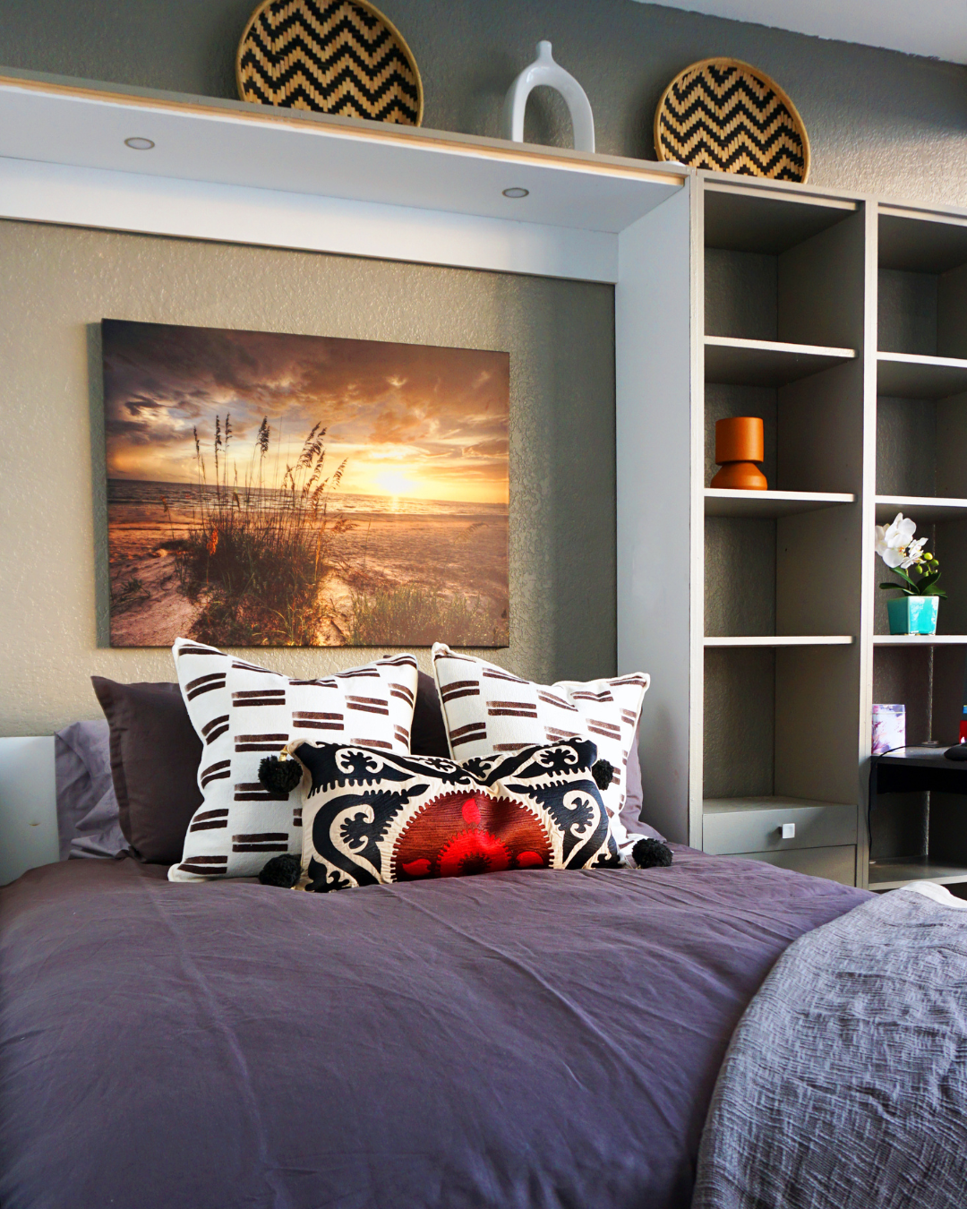 Custom Murphy bed with integrated shelving and desk, styled as a modern coastal guest bedroom with layered bedding, decorative pillows, and beach-inspired artwork