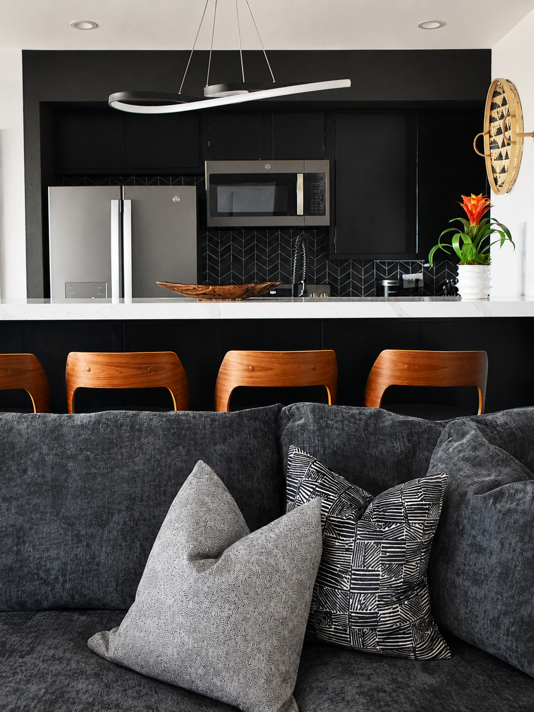 A modern black kitchen with a white marble countertop, wooden barstools, and a dark grey sofa in the foreground