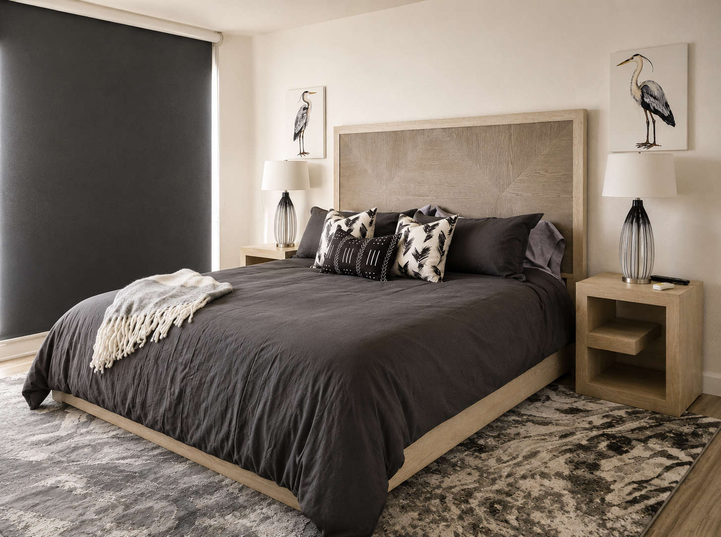 Modern coastal bedroom with a light wood platform bed, charcoal bedding, black and white accent pillows, matching nightstands, heron artwork, and contemporary neutral décor