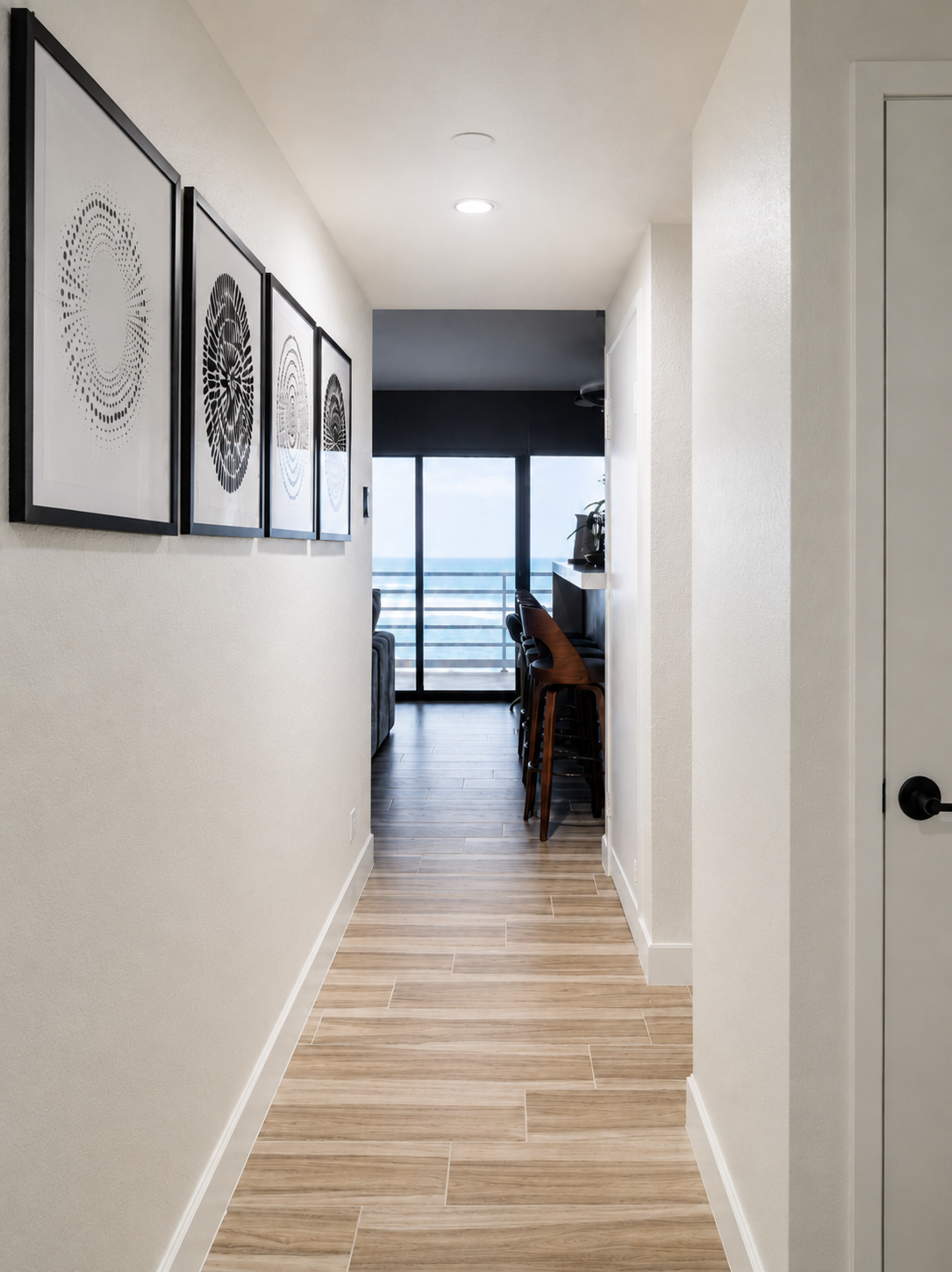 Modern coastal condo hallway with black-framed abstract artwork leading to an open living area, bar seating, and ocean-view balcony through floor-to-ceiling glass doors