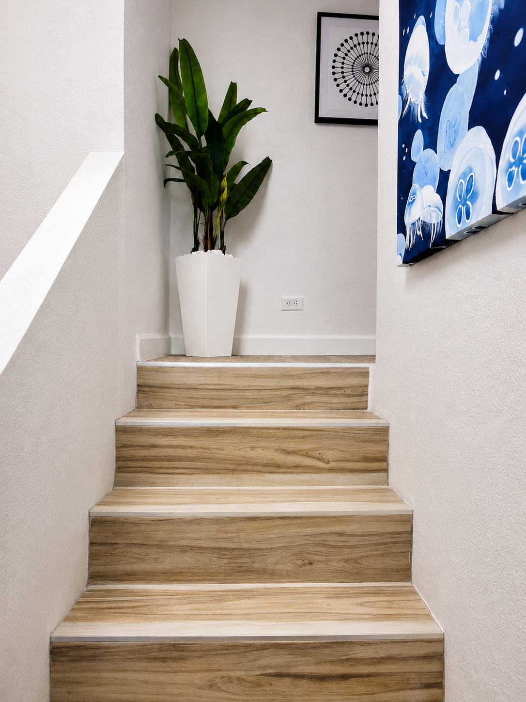 Modern coastal condo staircase with light blond wood-look stairs, white walls, oversized botanical planter, and blue jellyfish artwork in a bright, contemporary interior